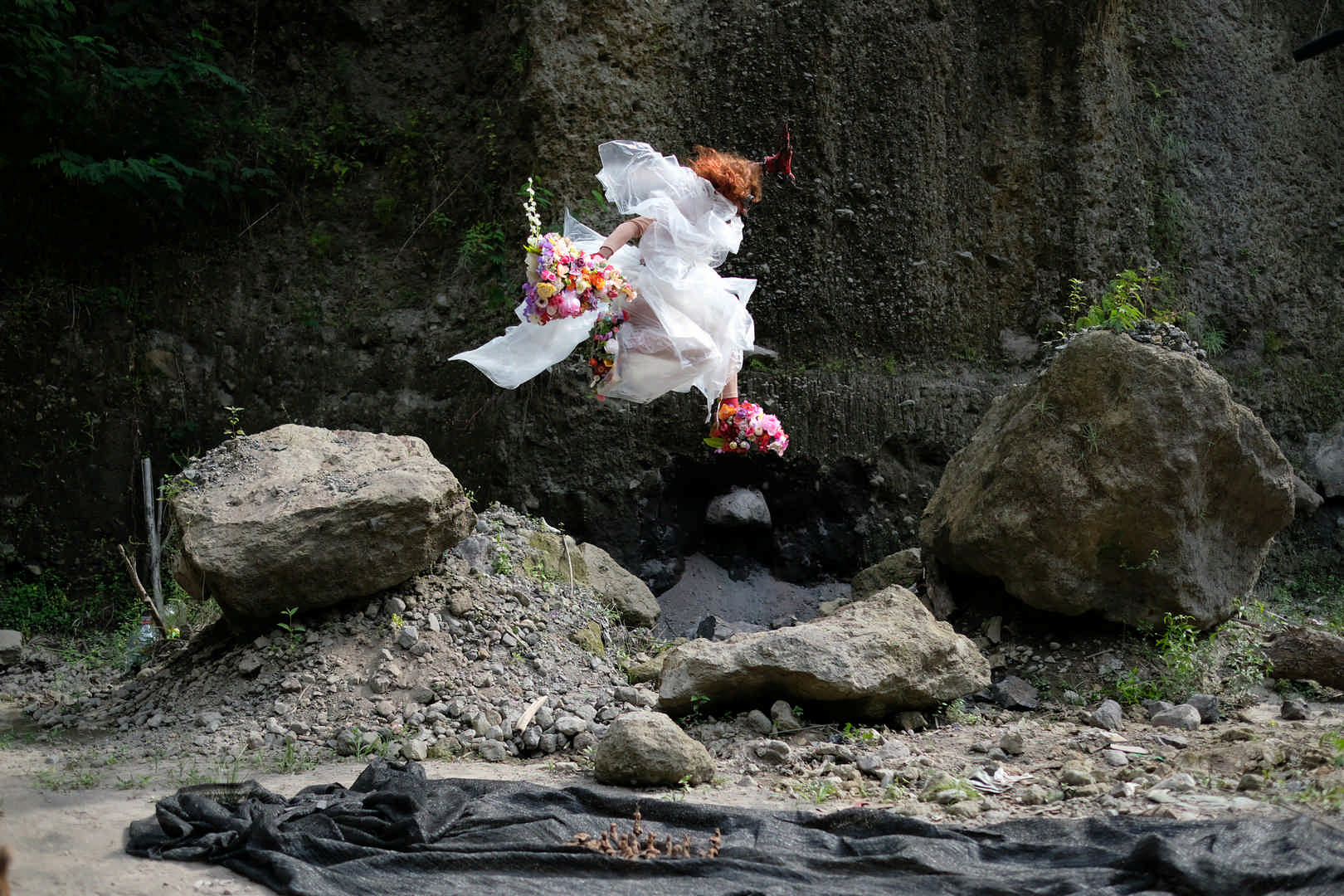 A person dressed in a big white dress, wearing a wig of long curly red hair, aviator goggles, and carrying enormous colourful bouquets of flowers is leaping high into the air from a swathe of grey cloth that is draped on the ground. There is a natural rock wall behind them, large boulders and a mound of broken down stone in their vicinity.