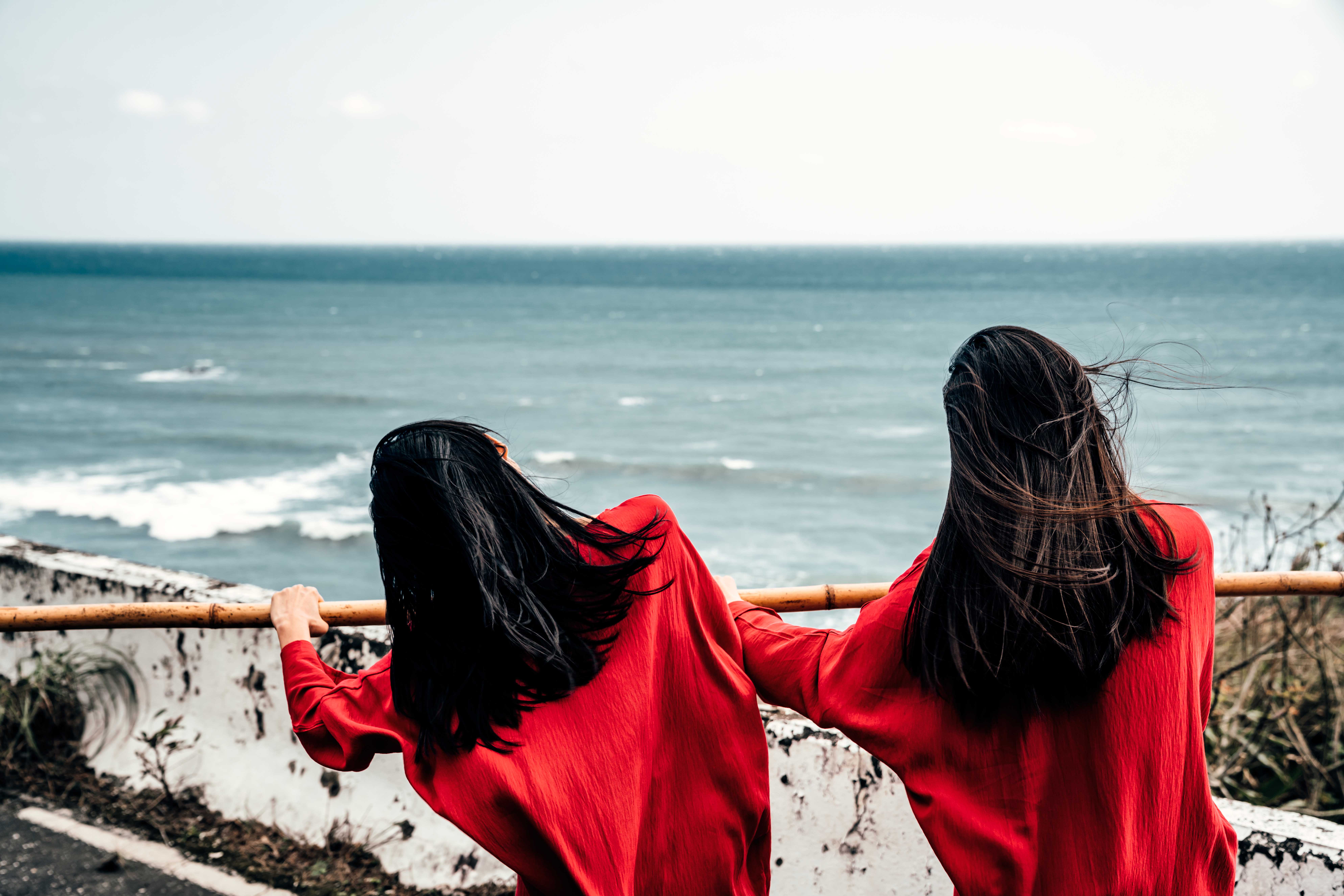 Two people with long black hair blowing in a sea breeze, their backs to the camera, both dressed in vibrant red, both holding onto a long piece of bamboo that dissects the whole frame. In the background, a white concrete wall and the sea.