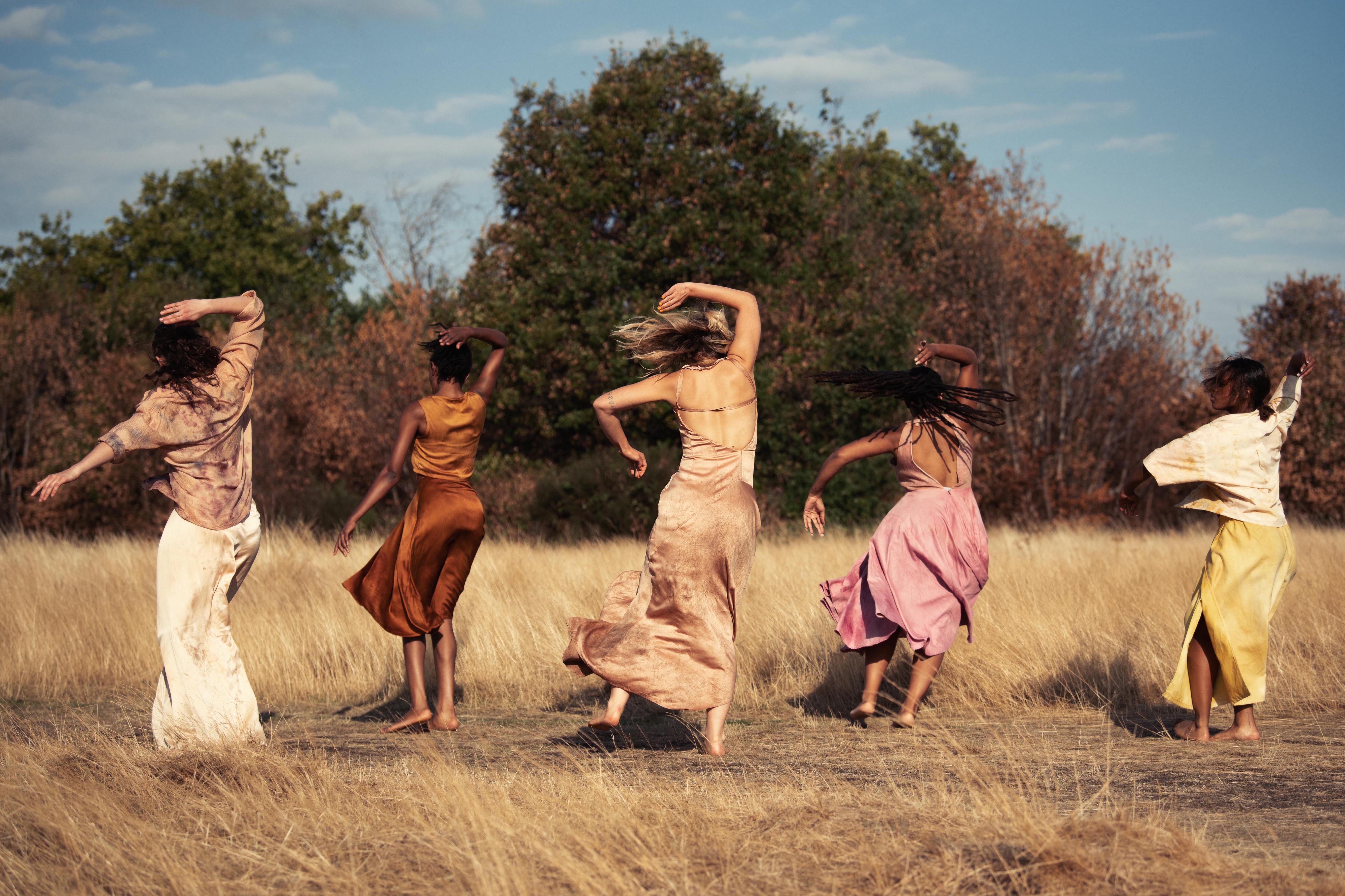 Five people are dancing in the middle of a field with long dry grass and trees behind. They all have their backs to the camera, all have their left arms held out from their bodies and bent at the elbow and right arm held out and raised above their head, their hair is swishing, the dance move looks energetic. They are dressed in shades of brown, pink, beige and orange.