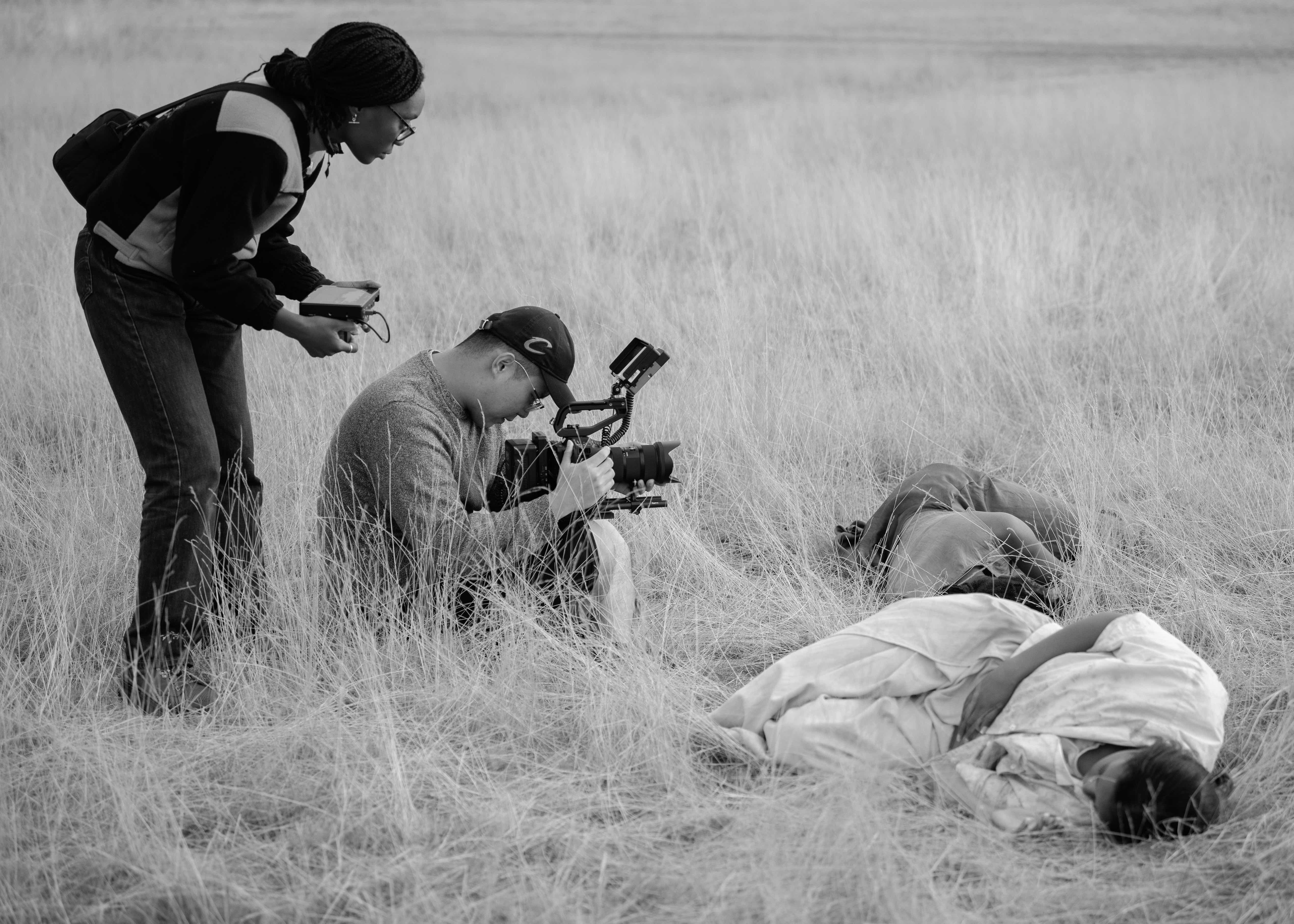 A photo in grayscale. There are two people lying amongst dry grass. There is another person sat on the ground filming these two people. A fourth person crouches over the person filming, watching through the lens of the camera.
