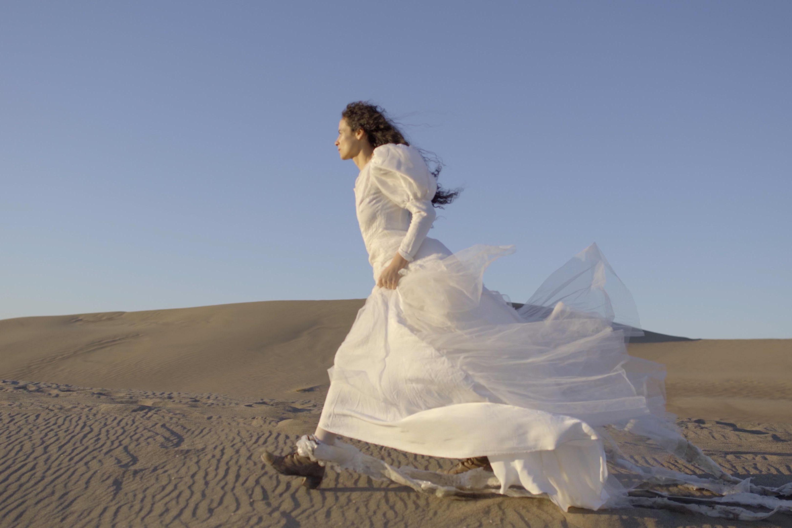 A person with long curly black hair strides through the desert wearing a white dress and brown heeled boots. Strips of white material are tied around their ankles and drag behind them as they walk.